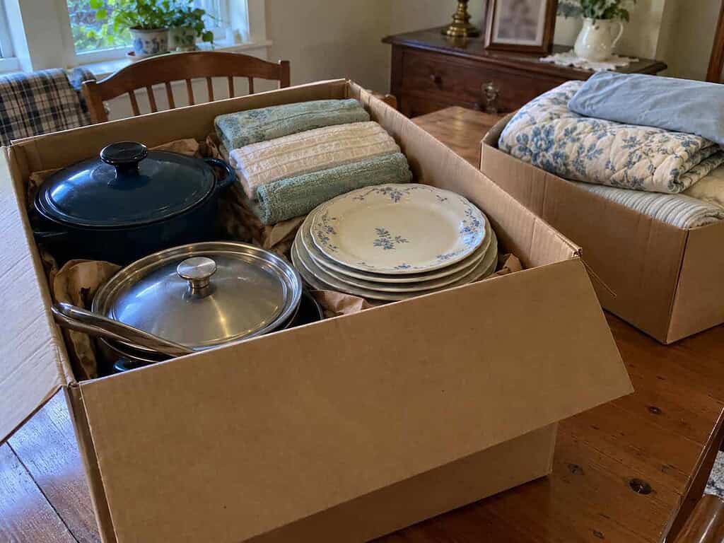 A cardboard box packed with clean pots, folded towels, and plates on a kitchen table, with a second box of bedding beside it.