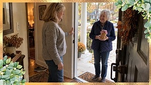 Smiling woman in a gray sweater holding her front door wide open for a white-haired friend in a navy vest who brings a jar of homemade applesauce on an autumn porch