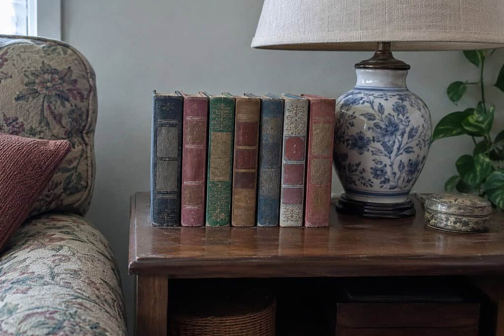 A short row of old colorful books on a living room end table beside a blue and white lamp