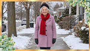 Smiling woman over 70 with short white hair in a lilac wool coat, red knit scarf, black pom beanie and leather gloves on a snowy small-town sidewalk