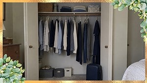 An open bedroom closet with a few navy, gray, cream and light blue clothes spaced on hangers and a small navy carry-on suitcase in front