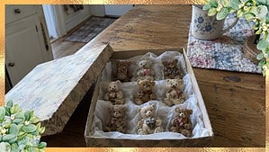 Box lined with white tissue paper holding nine small teddy bear figurines on a kitchen table, with the lid beside it