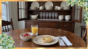 Breakfast on a rose-pattern china plate in front of a dining room hutch holding a gold-rimmed wedding china set and crystal glasses
