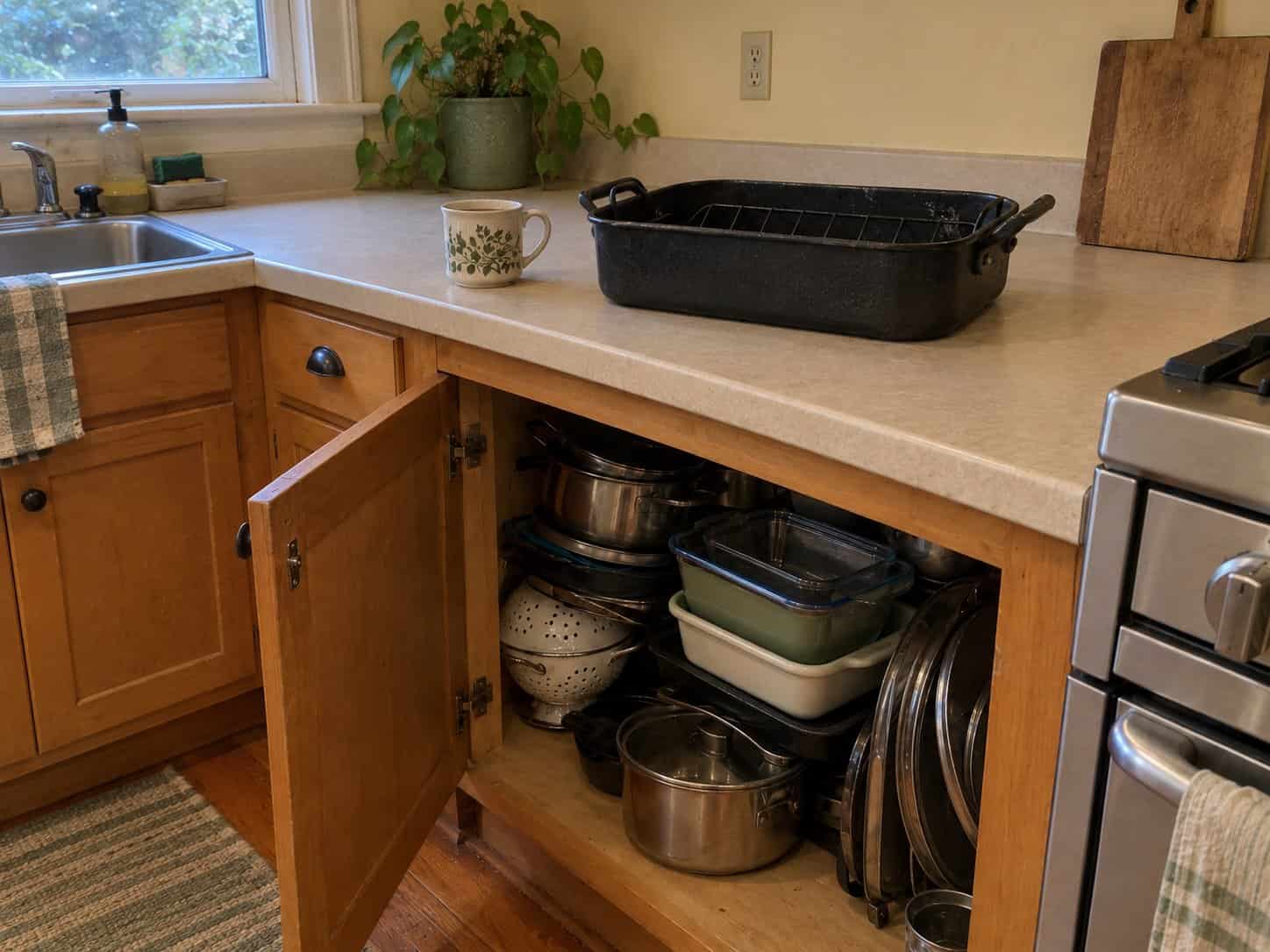 A heavy black roasting pan on a kitchen counter above an open cabinet crowded with pots, baking dishes and lids
