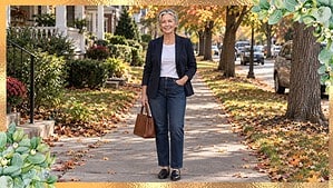 Smiling woman in her 60s in a navy blazer, white tee and dark jeans on a tree-lined sidewalk in fall