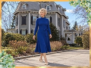 Smiling older woman with short platinum hair in a sapphire blue sweater dress and nude heels walking past a historic Newport house