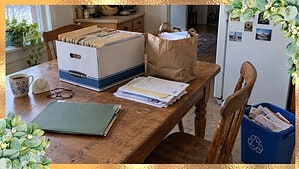 Kitchen table set up for sorting old papers with a Bankers Box of files, a paper bag for shredding, a green keep folder, a kitchen timer, and a recycling bin by the chair