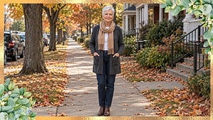 Smiling woman in her late 60s with a short white pixie cut in a long gray cardigan, camel scarf and dark jeans on a fall sidewalk