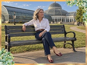 Smiling older woman with silver hair in a white shirt, dark jeans and red flats sitting on a bench in front of a glass conservatory