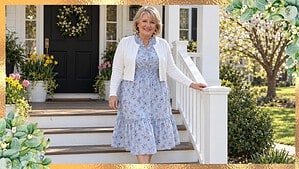 Woman in her 60s with a silver-blonde bob walking down sunny porch steps in a light blue floral midi dress, white cropped cardigan, and nude block-heel pumps