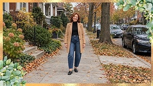 Woman over 60 in dark barrel jeans, a black turtleneck, an open camel coat and black loafers on a leafy sidewalk in the fall