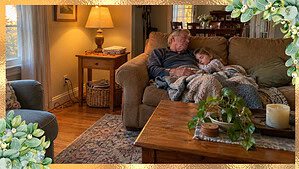 A grandfather and young grandchild asleep together on a living room couch, with a cluttered end table beside them