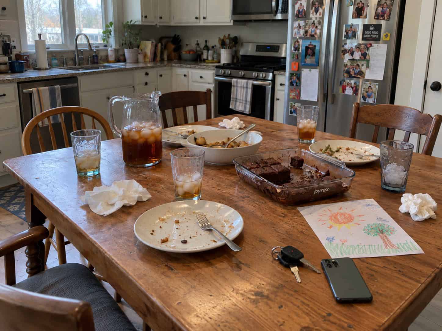 Wooden kitchen table right after Sunday dinner with empty plates, a pitcher of iced tea, half-full glasses, leftover brownies in a Pyrex dish, car keys, a phone, and a child's crayon drawing