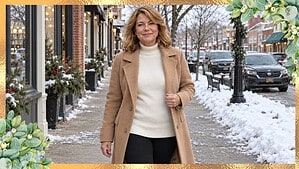 Woman in her 50s walking on a snowy small-town sidewalk in a camel wool coat, ivory turtleneck, and black leggings