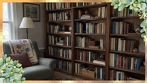 A wall of wooden bookshelves full of books in a spare room, with a quilt-covered reading chair and a floor lamp by the window