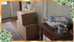 Plain moving boxes in a front hall beside a small table with a folded handmade quilt and a cup of coffee