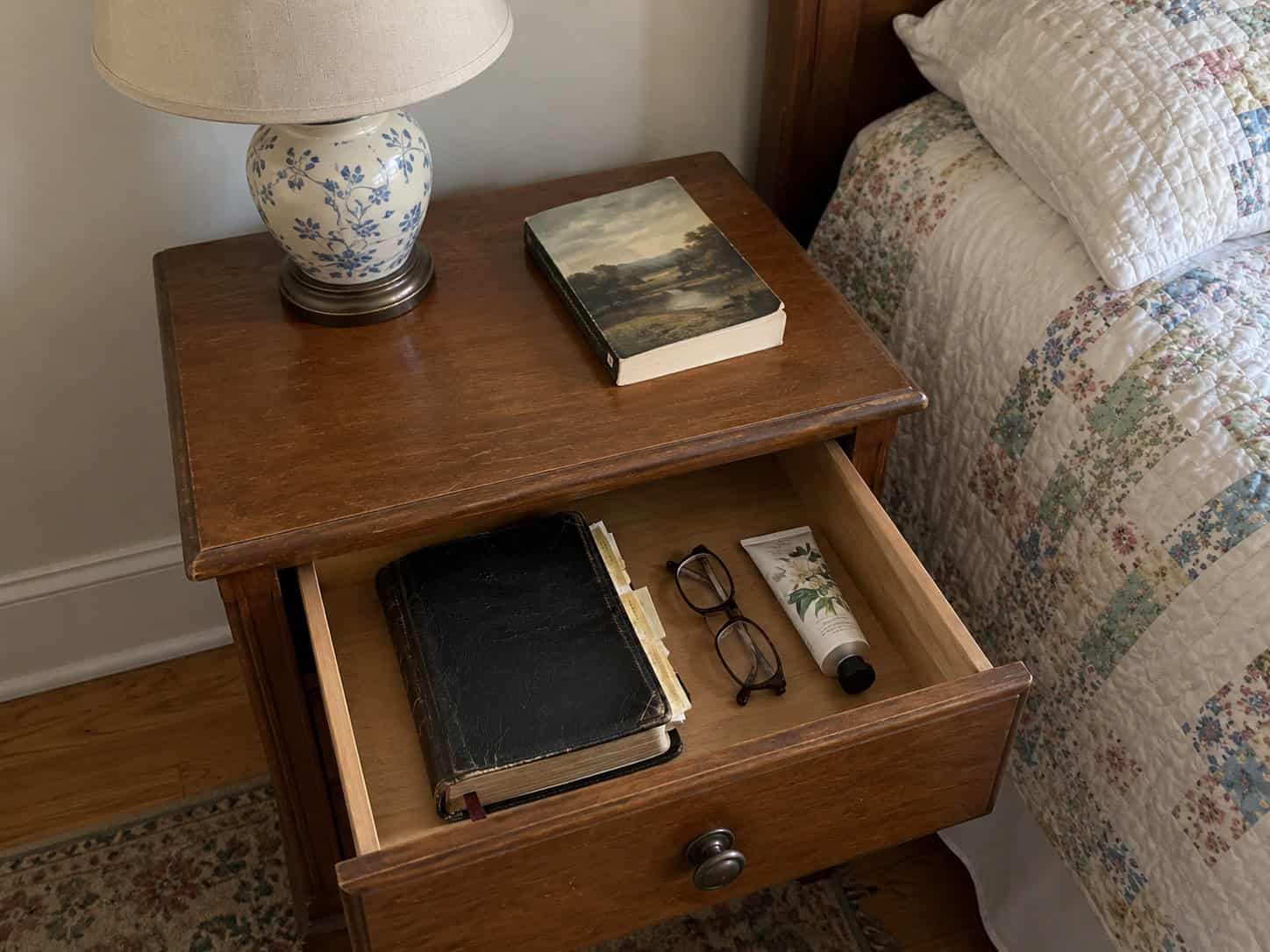 Well-worn leather Bible with reading glasses in an open nightstand drawer beside a quilted bed
