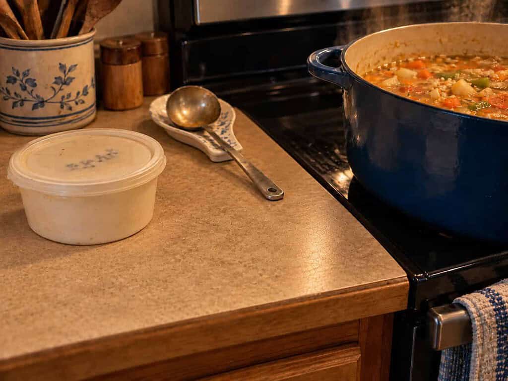 An old, well-used white margarine tub with faded handwriting on the lid, sitting beside a pot of vegetable soup