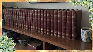 A long row of old red encyclopedia volumes with worn gold bands on a wooden shelf next to a cup of coffee