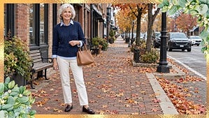 Smiling woman over 60 in a navy cable sweater over a blue oxford shirt, cream pants, brown loafers and a tan handbag on a leafy fall sidewalk