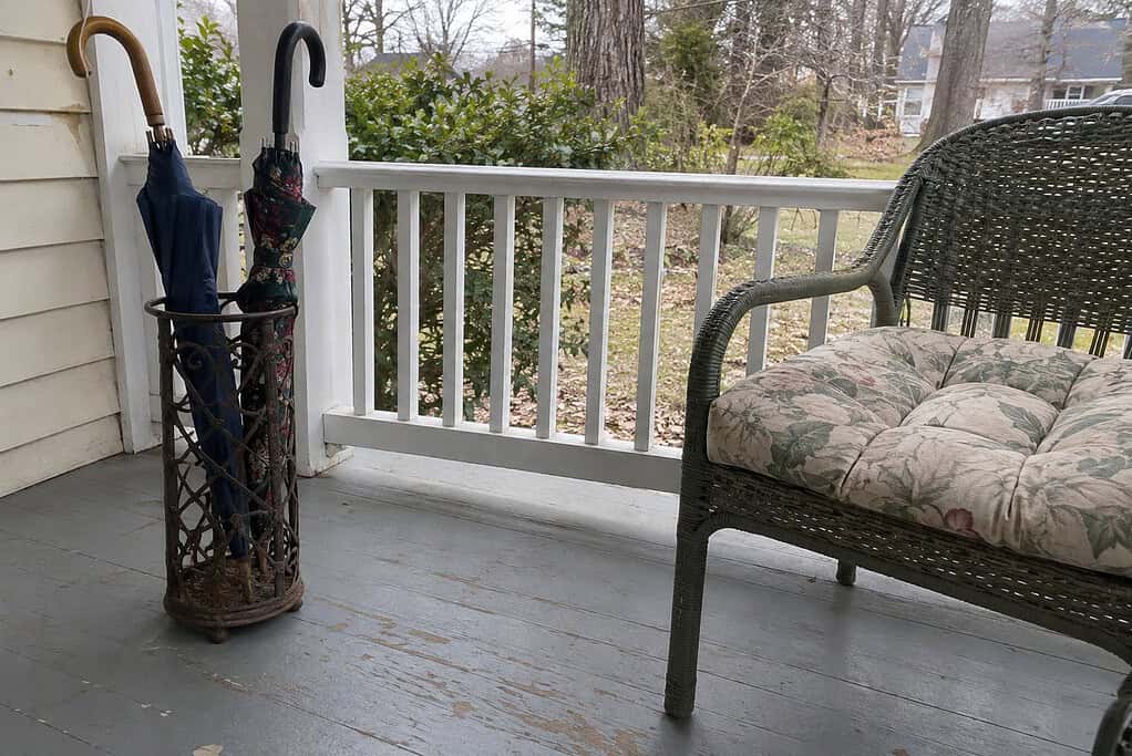 An old metal umbrella stand with two umbrellas on a front porch beside a wicker chair