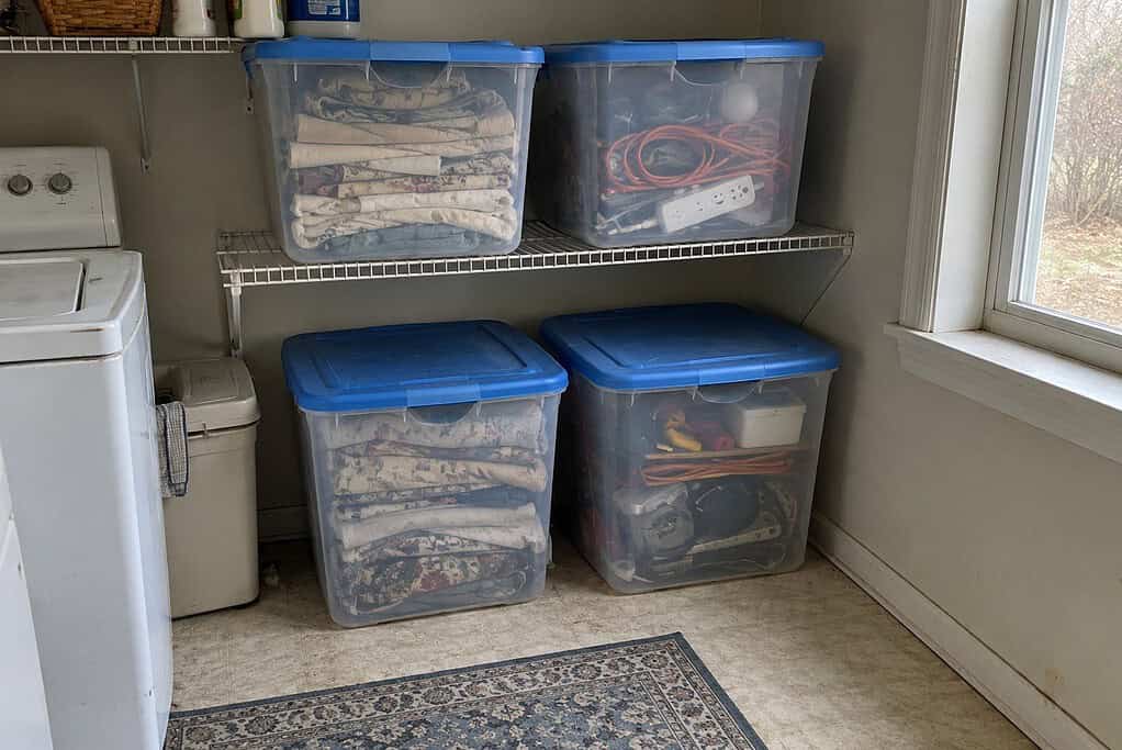 Four clear plastic bins with blue lids full of old curtains and cords in a laundry room