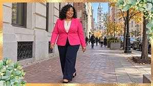Plus size woman in a hot pink blazer and navy wide-leg trousers walking on a Philadelphia sidewalk with City Hall behind her