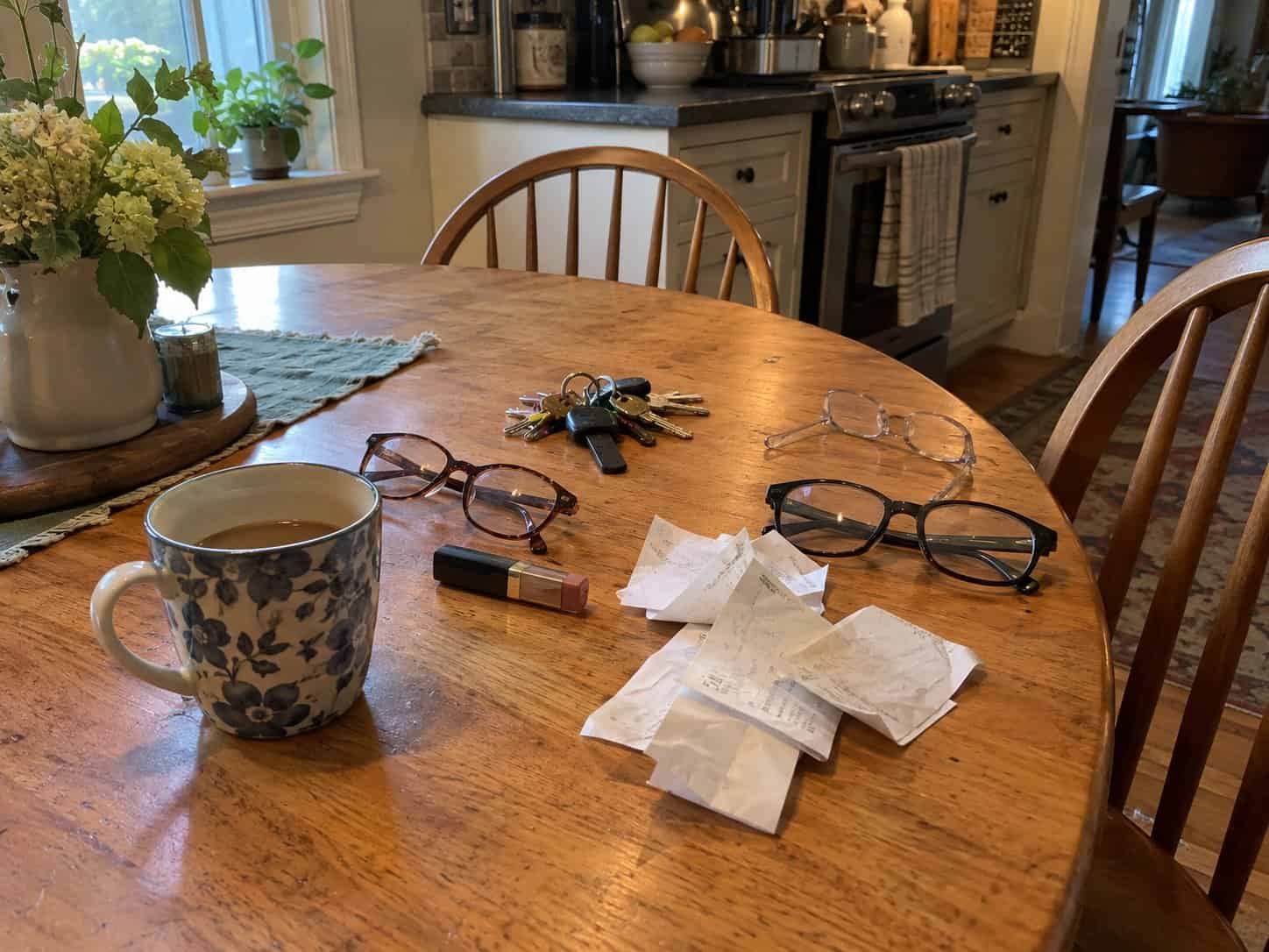 Reading glasses, a ring of keys, crumpled receipts, a lipstick and a coffee mug on a wooden kitchen table