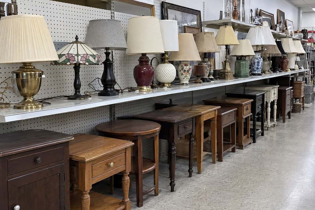 A thrift store shelf full of used table lamps above a row of wooden end tables