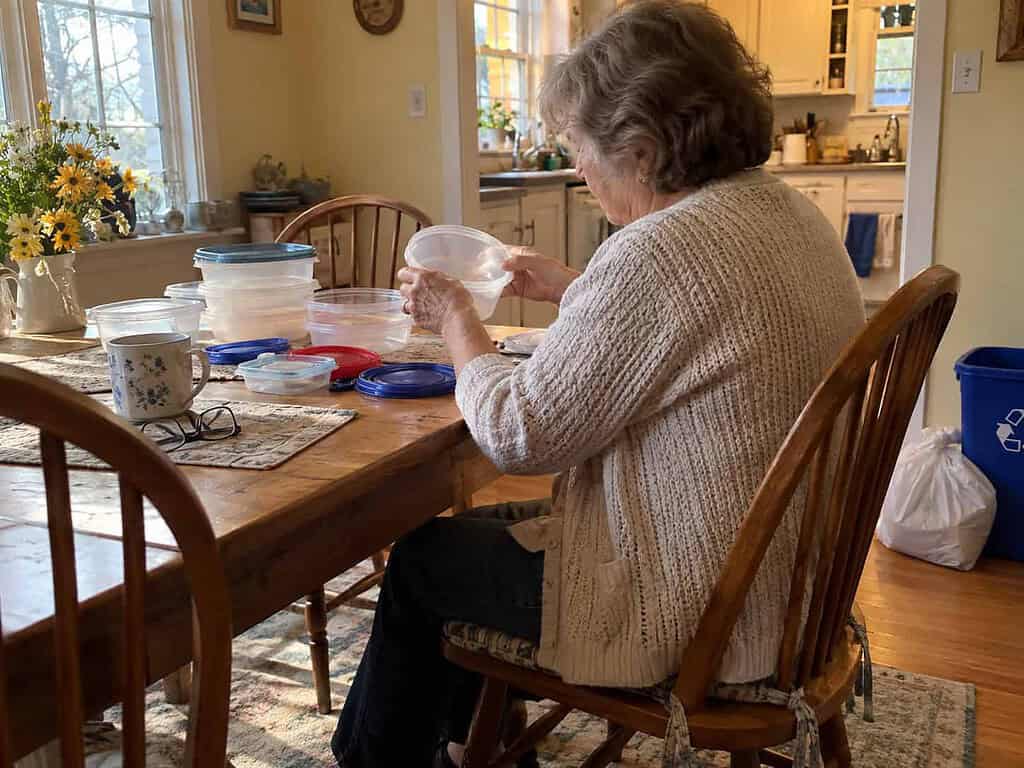 A woman in her 60s sitting at her kitchen table sorting plastic food containers, with a trash bag and recycling bin beside her chair