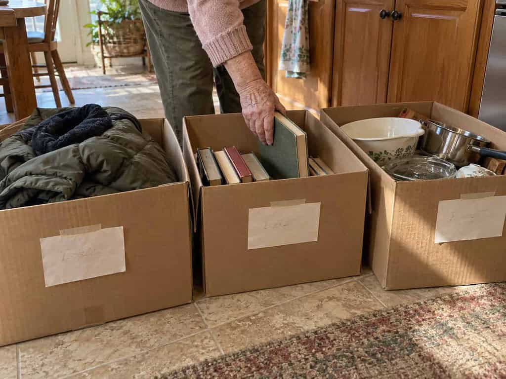 Three cardboard boxes on a kitchen floor holding a coat, books, and kitchen items, with a woman's hand placing a book in one.