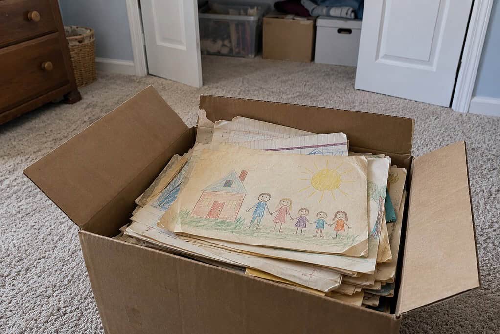 Open cardboard box of old school papers on a spare room carpet, with a child's crayon drawing of a house, a sun and five stick people on top