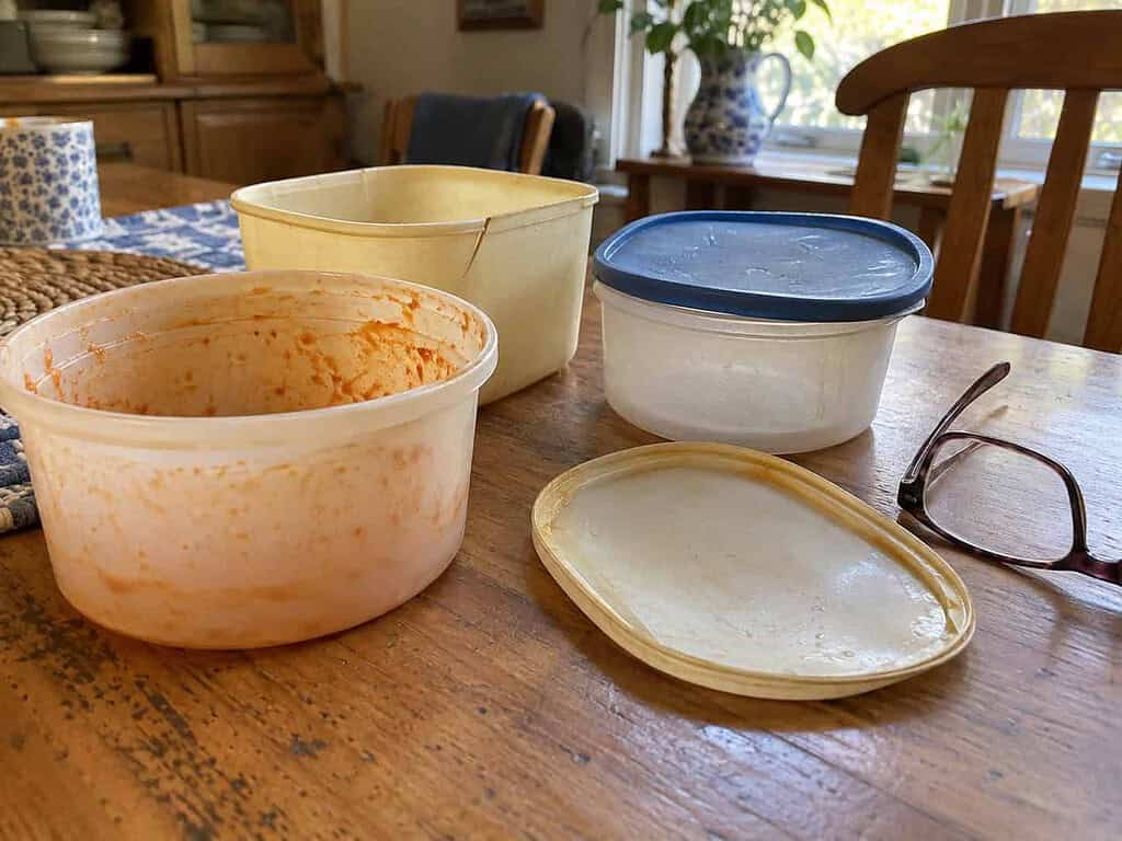 Old plastic food containers with a tomato sauce stain, a cracked rim, and a bent lid, set aside beside reading glasses