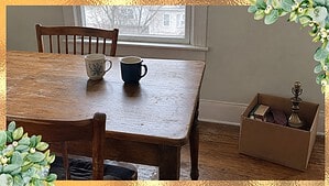Two coffee mugs on a wooden kitchen table by a window, with a box of books, a tie and an old lamp on the floor to give away