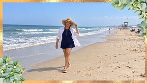 Woman over 70 in a navy swim dress, white linen shirt, straw sun hat and tan sandals walking on the beach