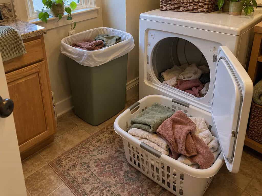 A tall trash can lined with a bag and holding donated clothes in the corner of a laundry room.