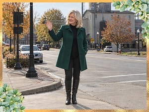 Smiling older woman with blonde hair in an emerald green A-line coat, black mock-neck sweater, black jeans and knee-high boots waving on a small-town street in fall