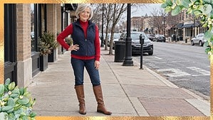 Woman over 50 in a red turtleneck, navy quilted vest, dark jeans and brown riding boots on a small-town main street