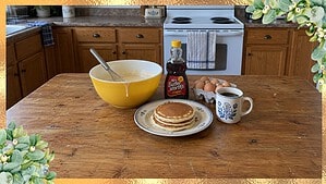 Yellow vintage Pyrex mixing bowl with pancake batter next to a stack of pancakes and syrup on a kitchen table