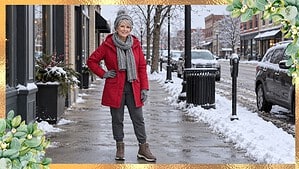 Woman over 60 in a red parka, gray knit hat, scarf and gloves, gray joggers and hiking boots on a snowy small-town main street