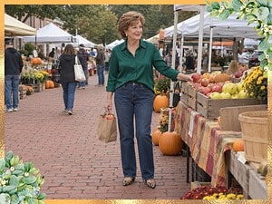 Older woman in an emerald green satin blouse with a French tuck, dark jeans and leopard flats reaching for apples at a fall farmers market
