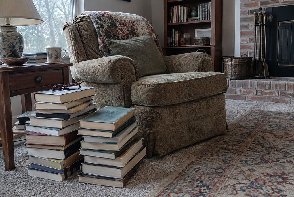 Tall stacks of books on the floor beside an armchair, with reading glasses on top of one stack