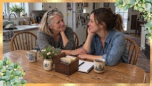 Mother in a gray sweater and her grown daughter in a denim shirt talking and smiling at a wooden kitchen table with coffee mugs, a notebook, and an old wooden recipe box