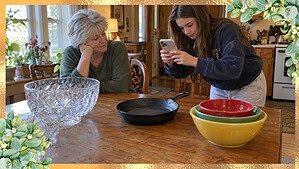A woman in her 60s watches her teenage granddaughter photograph a cast-iron skillet on a kitchen table