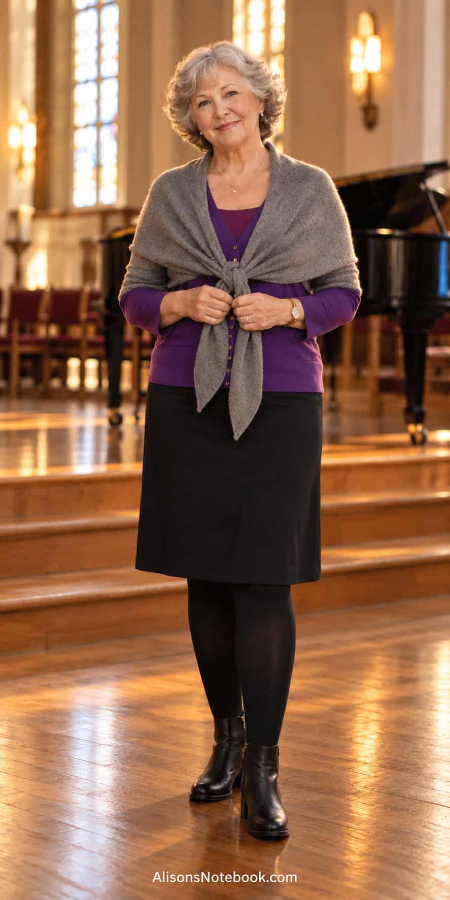 Woman around eighty with short curly silver hair wearing a purple cardigan, gray knit shawl, black knee-length skirt, black tights, and black ankle boots beside a piano in a church.