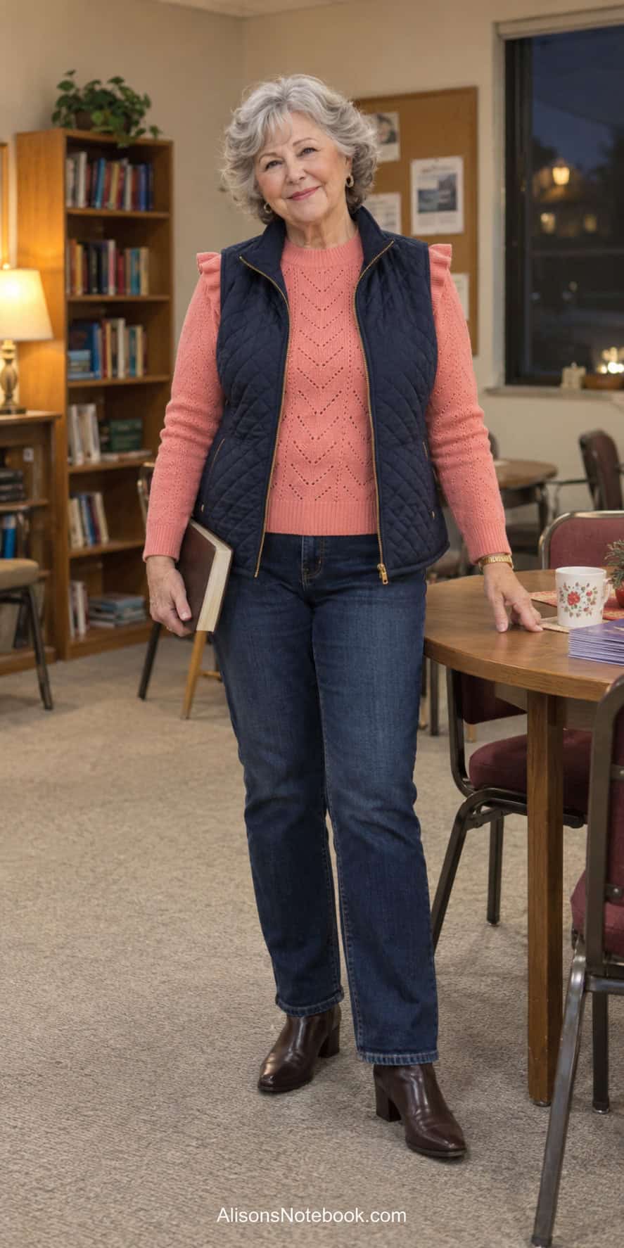 Woman around eighty with short curly silver hair wearing a coral knit sweater, navy quilted vest, dark straight jeans, and brown ankle boots in a church classroom.