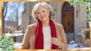 Woman around eighty with short curly silver hair wearing a cream sweater, an open camel coat, and a red scarf outside a stone church in winter.