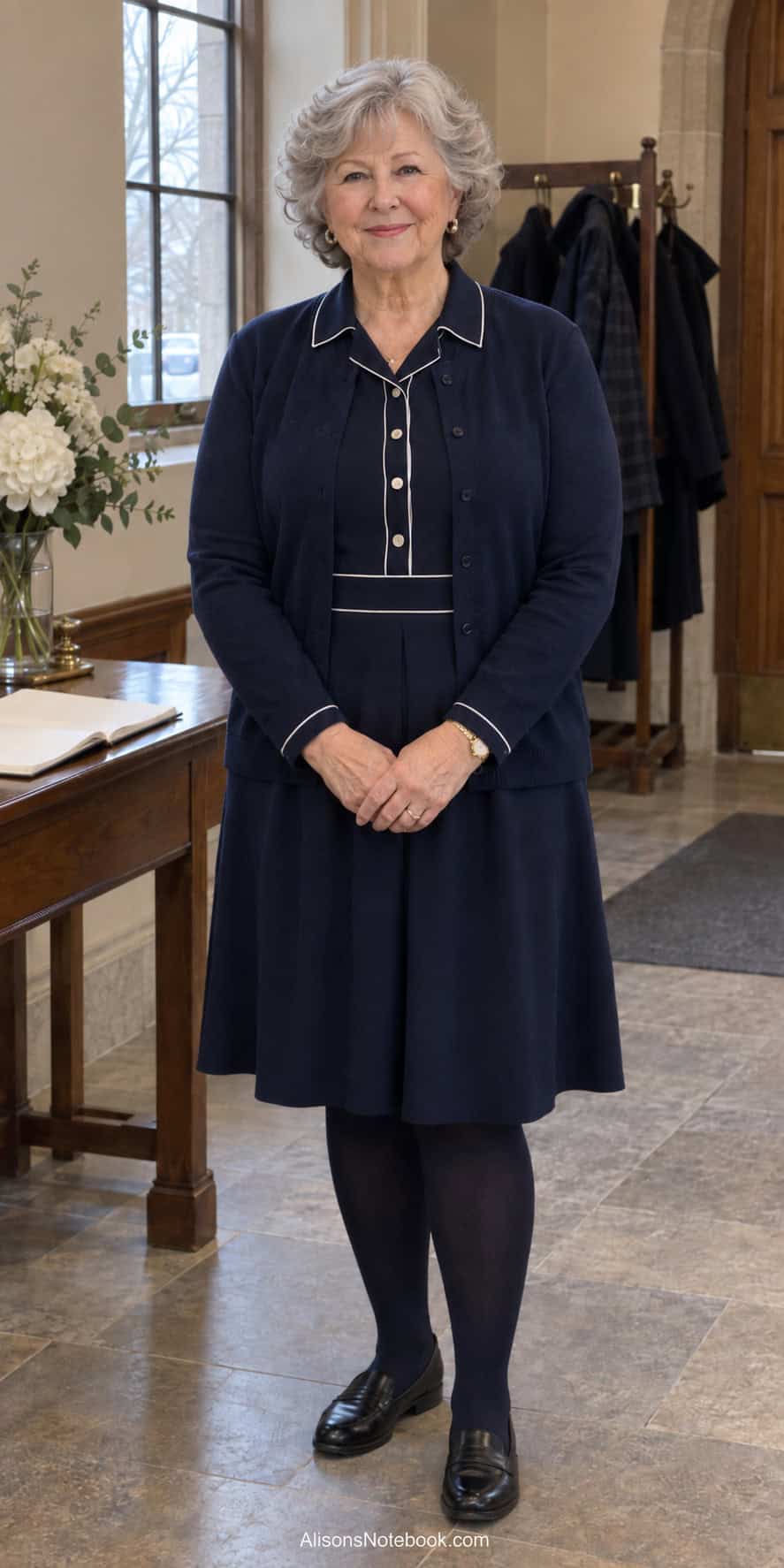 Woman around eighty with short curly silver hair wearing a navy collared knit dress, navy cardigan, navy tights, and black loafers in a church entry hall.