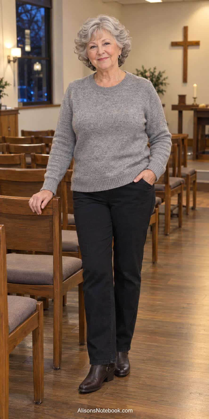 Woman around eighty with short curly silver hair wearing a heather gray sweater, dark straight-leg pants, silver hoop earrings, and brown ankle boots in a small church chapel.