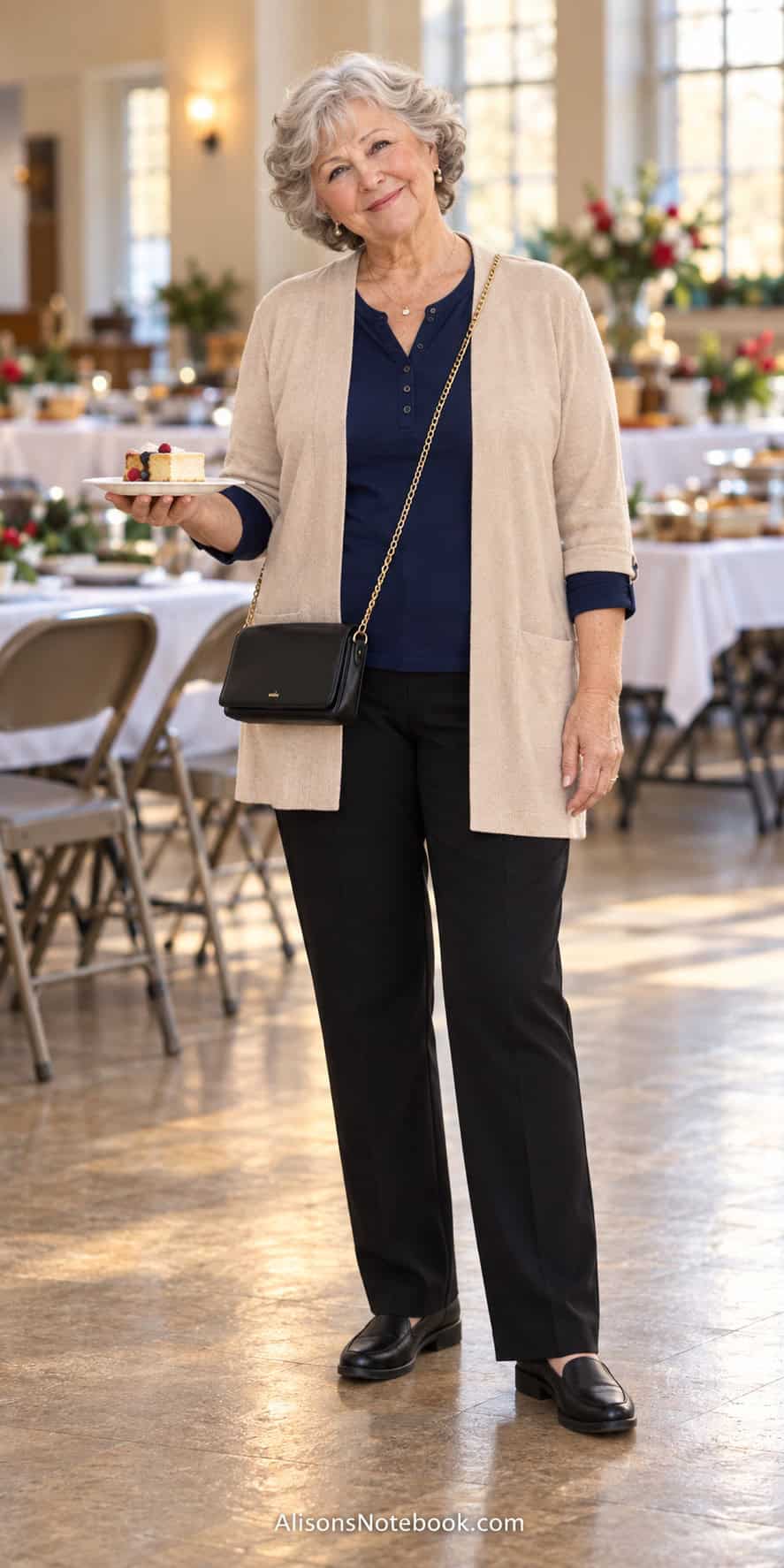 Woman around eighty with short curly silver hair wearing a navy top, oatmeal open cardigan, black pants, a black crossbody bag, and black slip-on shoes in a church fellowship hall.
